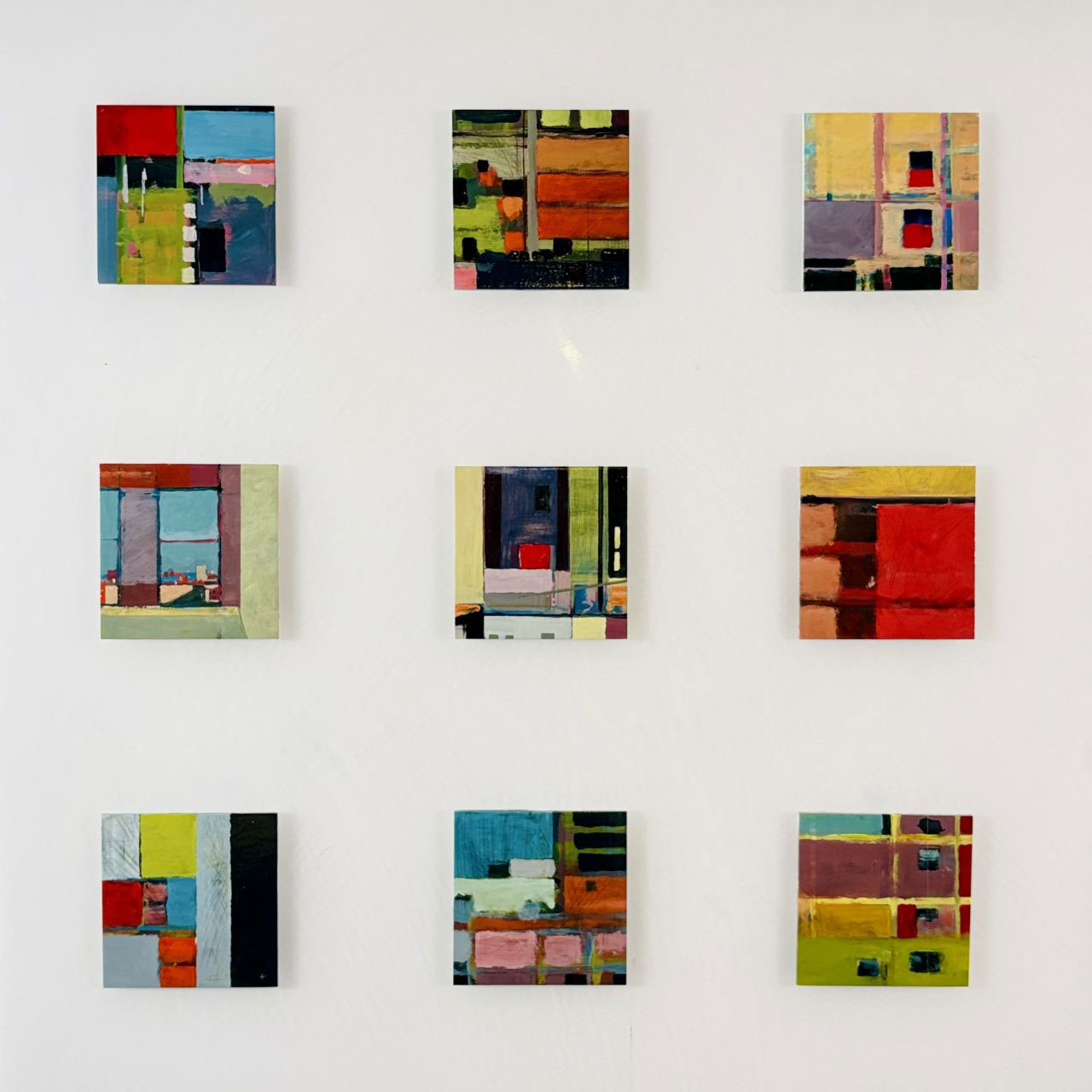Nine colorful abstract paintings by Barry Ginder in a three-by-three grid are displayed on a white wall, each featuring geometric shapes and varied color blocks.