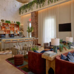 Modern hotel lounge designed by LEGEARD STUDIO, featuring a stylish bar, comfy sofas and armchairs, large windows with sheer curtains, and lush greenery decor on walls and shelves.