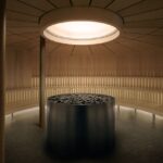 A modern circular sauna with wooden benches lining the walls, a central metal stove filled with rocks, and a gong hanging on the right. Natural light enters from a round ceiling window.
