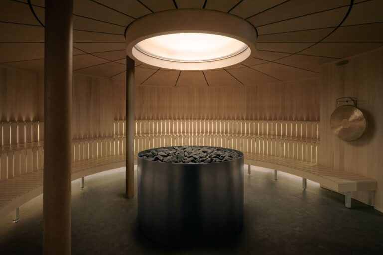 A modern circular sauna with wooden benches lining the walls, a central metal stove filled with rocks, and a gong hanging on the right. Natural light enters from a round ceiling window.