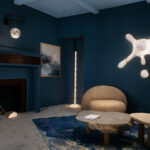 Modern living room with dark blue walls, abstract wall lights, a beige curved sofa, stone coffee tables, and a decorative rug. A basketball hoop light fixture hangs above the fireplace—a perfect Friday Five feature for design inspiration.
